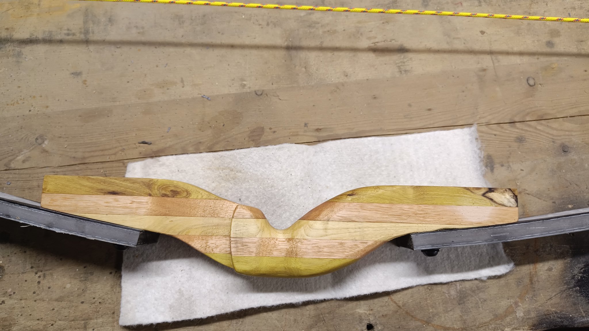 Vor uns auf dem Tisch liegt ein Griffstück eines Bogens, das aus verschieden farbenes Holz zusammengeleimt wurde. An ihm sind beide Wurfarme befestigt. Das Holz wurde geölt und glänzt ein wenig