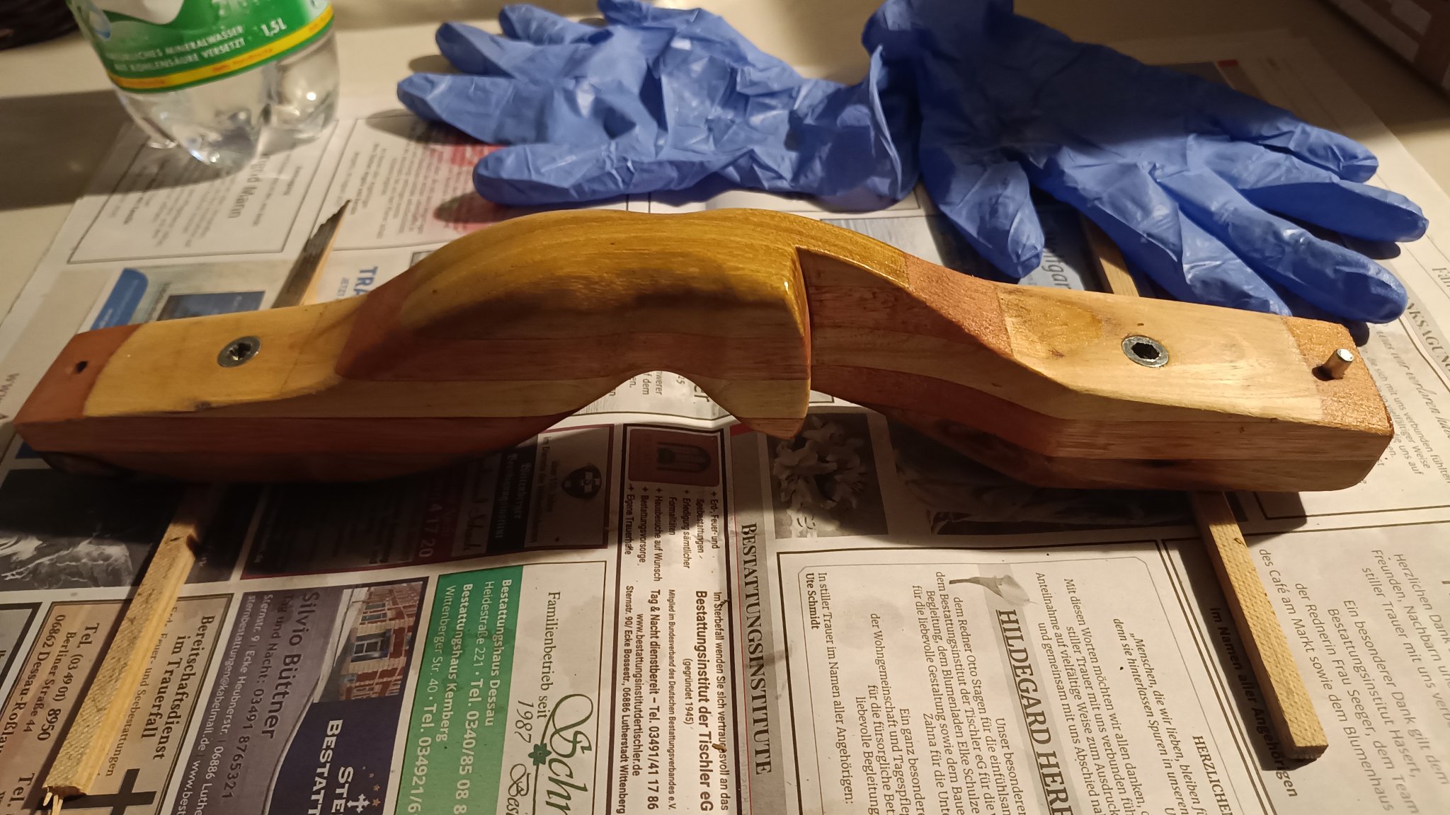 Vor uns auf dem Tisch liegt ein selbst gebautes Griffstück für einen Bogen. Das Holzstück wurde aus mehreren Lagen unterschiedlich farbigem Holz zusammen geleimt und ausgesägt und geschliffen. Hier hat es eine neue Ölschicht bekommen, das jetzt in das Holz einziehen soll