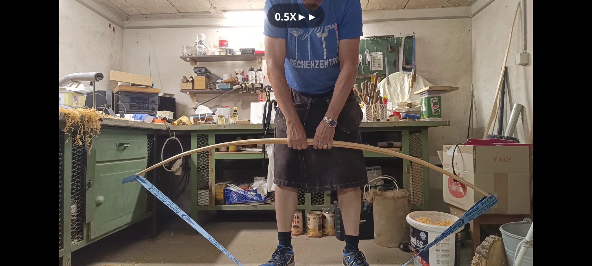 in einer ziemlich rumpligen Werkstatt steht ein Mann im Kilt vor uns. Er trägt ein blaues tShirt auf dem harken und Laubkratzen abgebildet sind und darunter steht: Rechenzentrum Mit beiden Händen zieht er an seinem Bogenrohling, das an beiden Enden mit einem Spannband verbunden ist. Dadurch sieht man, wie sich der Rohling langsam biegen lässt. Zusätzlich erkennt man Stellen, wo die Biegung nich nicht so gut aussieht